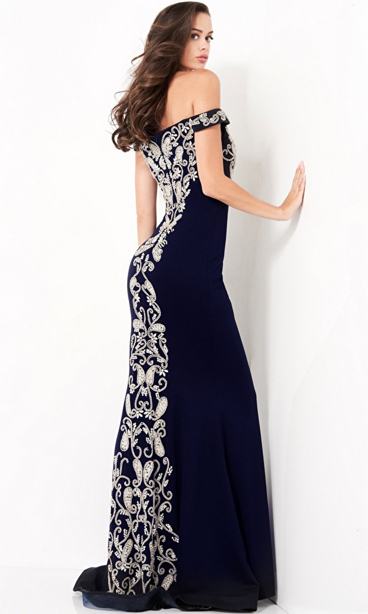 A woman stands facing a white wall, looking over her shoulder and touching the wall, wearing the Jovani Embroidered Off-Shoulder Prom Dress 02576 in navy blue with silver embroidered patterns.